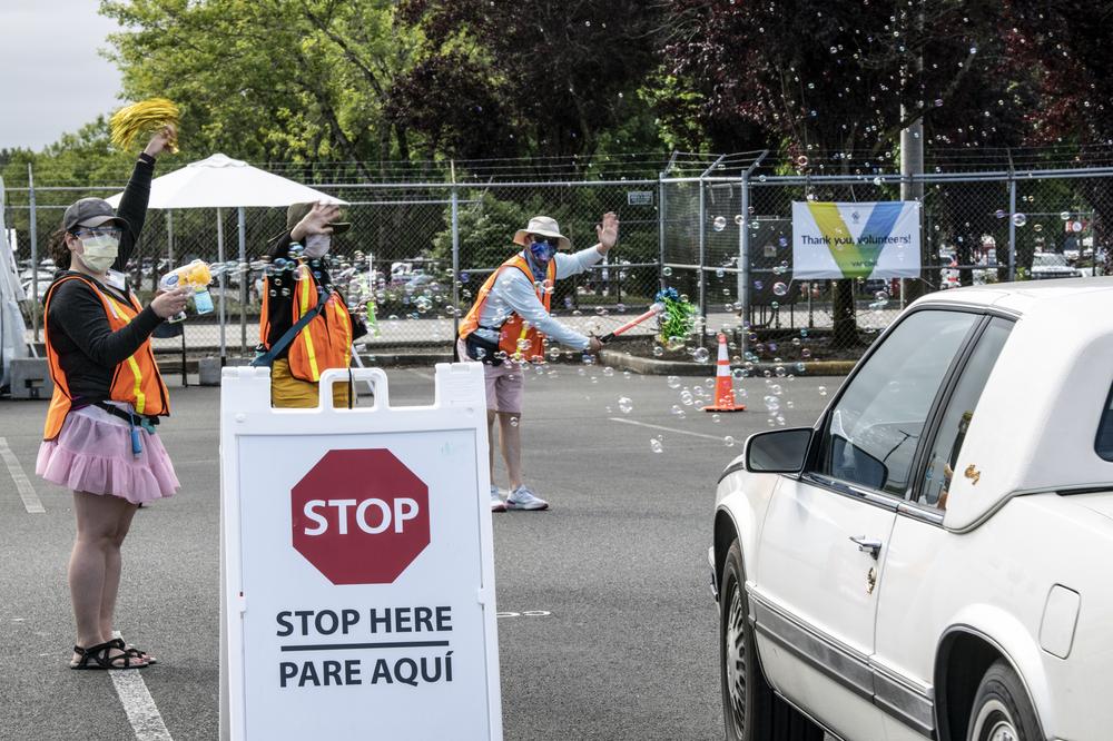 Three people with orange vests and pom poms wave at a white car, who is exiting the PDX Red Lot vaccine clinic